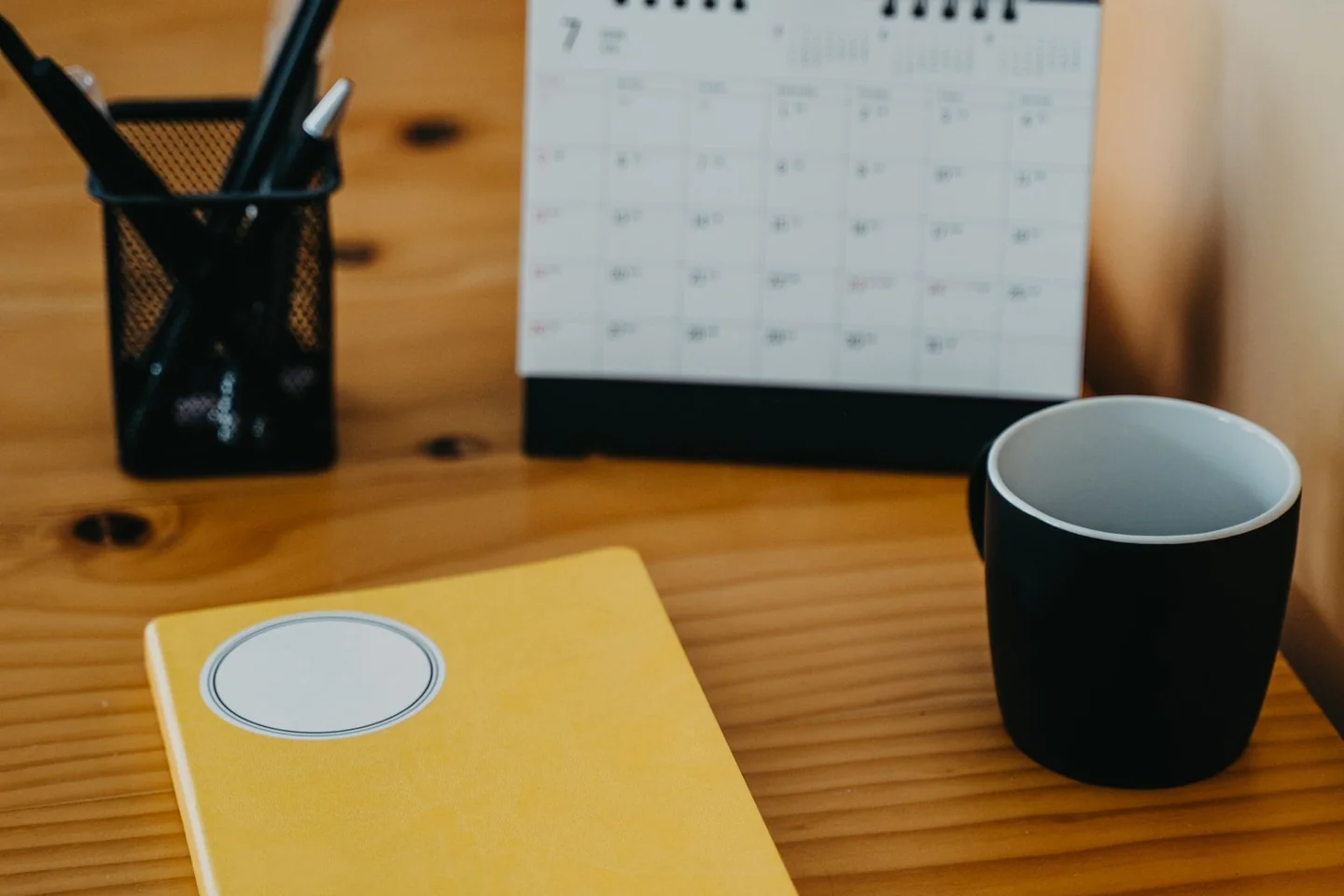 A calendar, notebook and mug on a wooden desk.